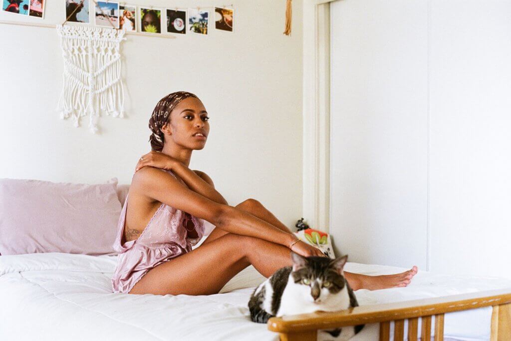 Image by Danielle Spires titled "Rikki"; a woman sits on a bed with a cat